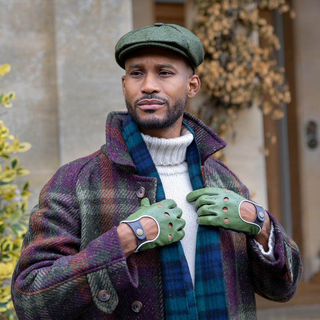 A man wearing a green tweed newsboy cap, a blue and green plaid scarf, and green leather driving gloves outdoors in front of a stone building.