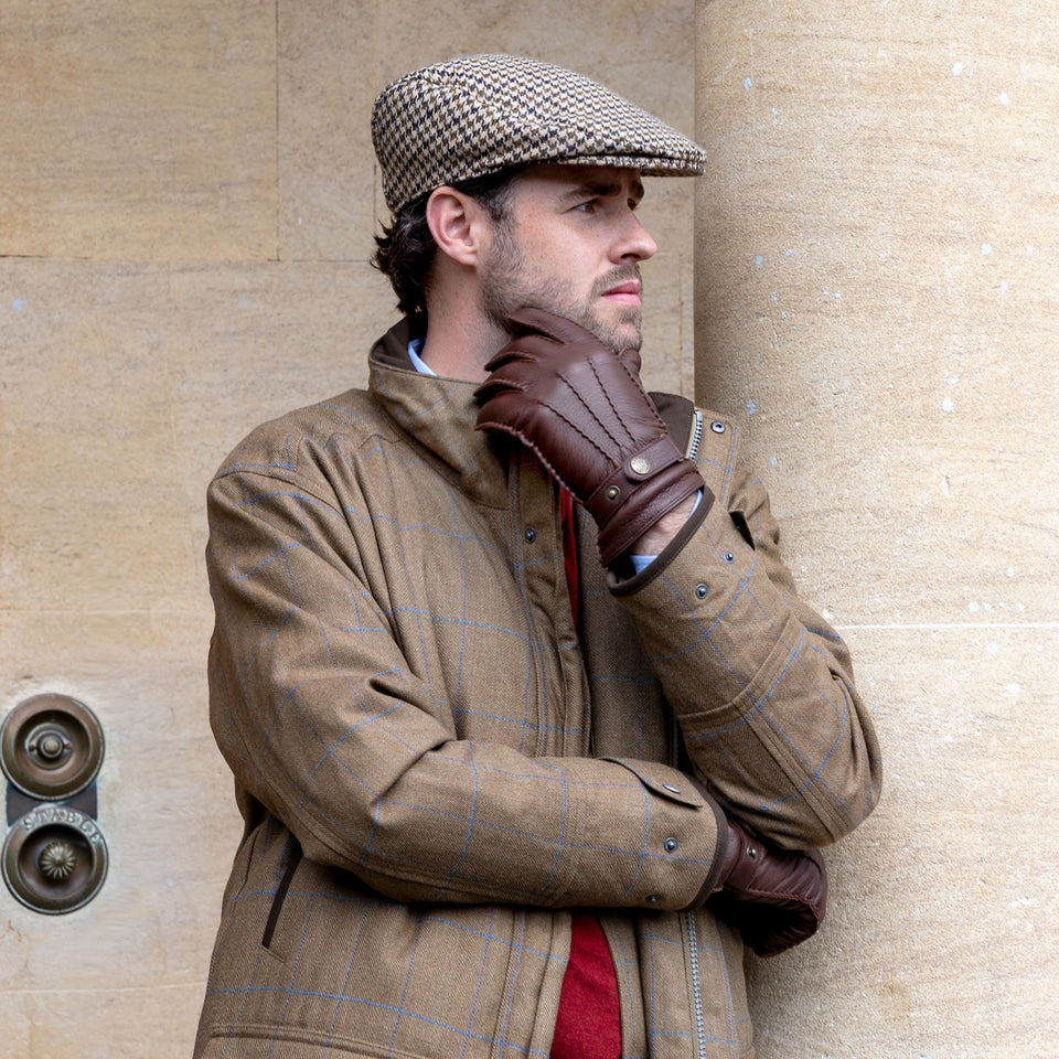 Man wearing brown leather gloves and a dogtooth check flat cap stood by a stone pillar