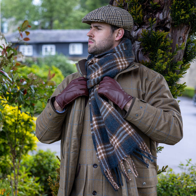 A man wearing a brown tweed jacket, a blue and brown plaid blanket scarf, brown leather gloves, and a brown dogtooth check flat cap, standing outdoors in front of a tree.