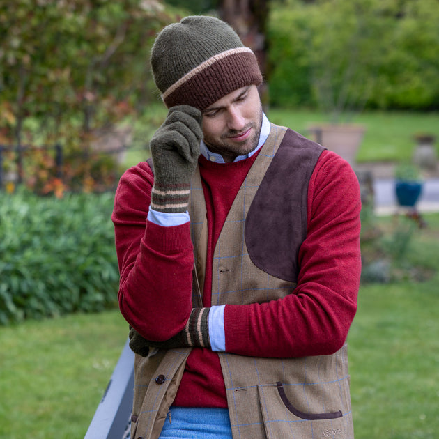A man wearing a green and brown striped knitted beanie hat and matching knitted gloves in a garden.