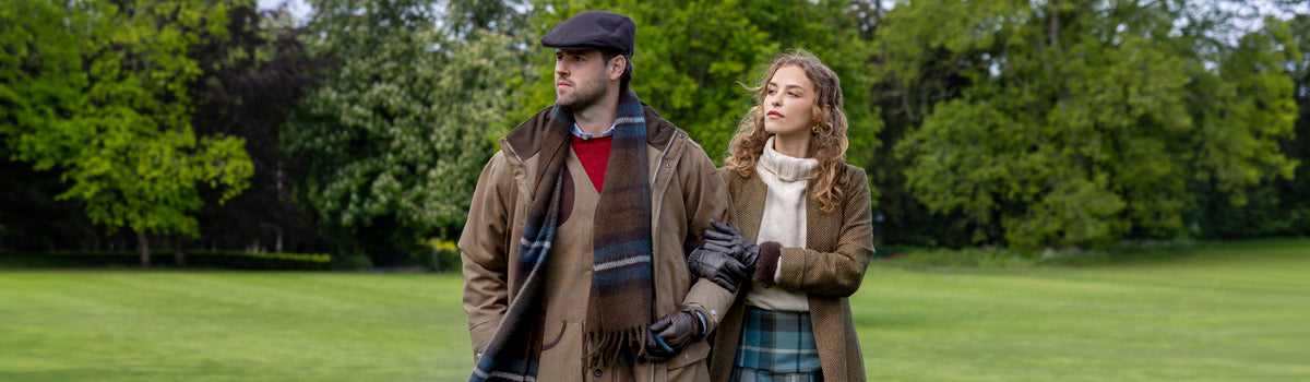 A man and a woman standing arm in arm in a park. The man wears brown leather gloves, a brown checked scarf, and a flat cap, while the woman wears brown faux fur-trimmed leather gloves.