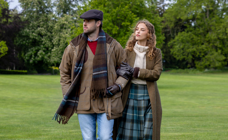 A man and a woman standing arm in arm in a park. The man wears brown leather gloves, a brown checked scarf, and a flat cap, while the woman wears brown faux fur-trimmed leather gloves.