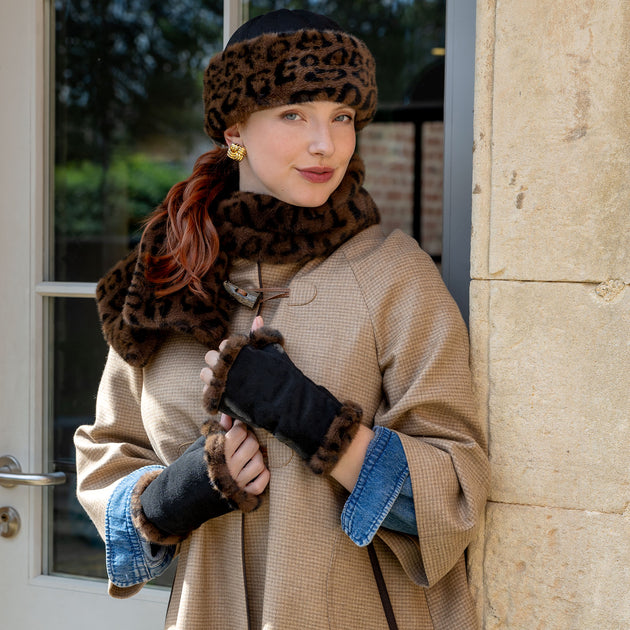 Woman wearing a animal print faux fur-trimmed hat, matching faux fur scarf and fingerless mittens stood by a door outside