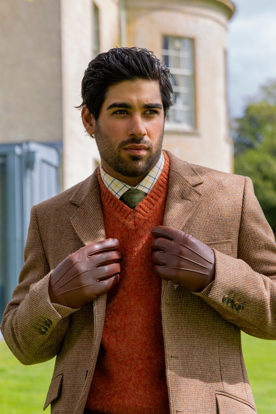Man wearing brown leather gloves, brown jacket and orange sweater in front of a building