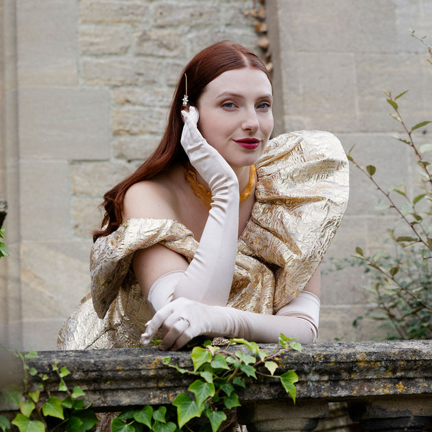 Woman wearing long satin gloves and a gold ballgown at a stone balcony