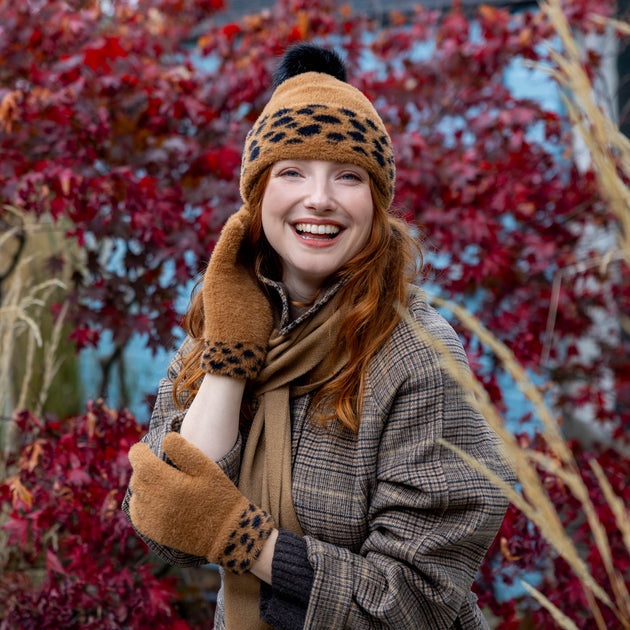 A woman outdoors wearing a tan and black leopard-print knit beanie with a pom-pom, matching knitted gloves, and a brown plaid coat, with red autumn foliage in the background.