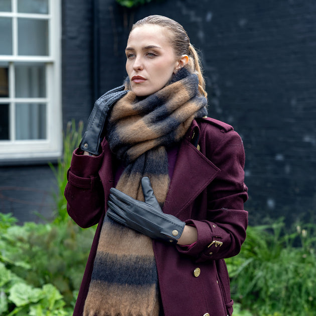 A woman wearing a burgundy coat, a brown and black striped blanket scarf, and black leather gloves, standing outdoors in front of a dark brick wall.