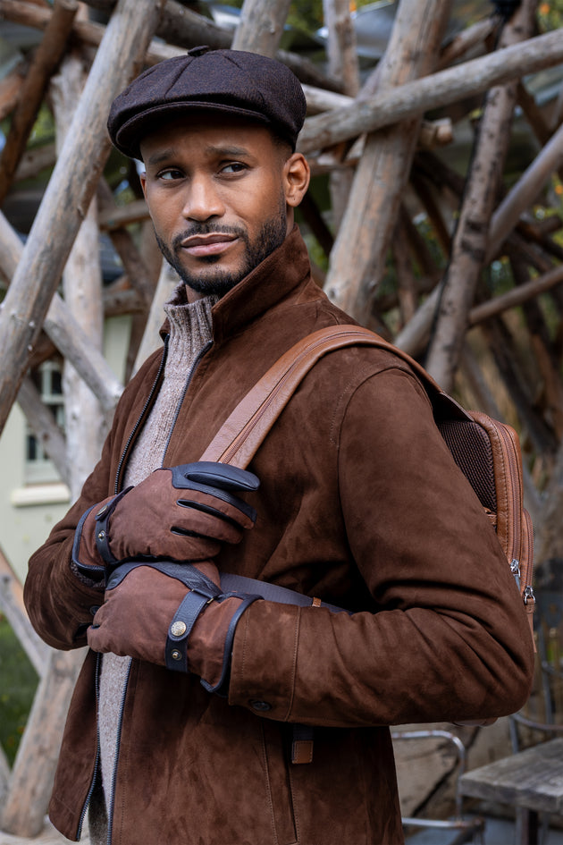 Man wearing a brown tweed newsboy cap and waxed cotton gloves carrying a leather packpack
