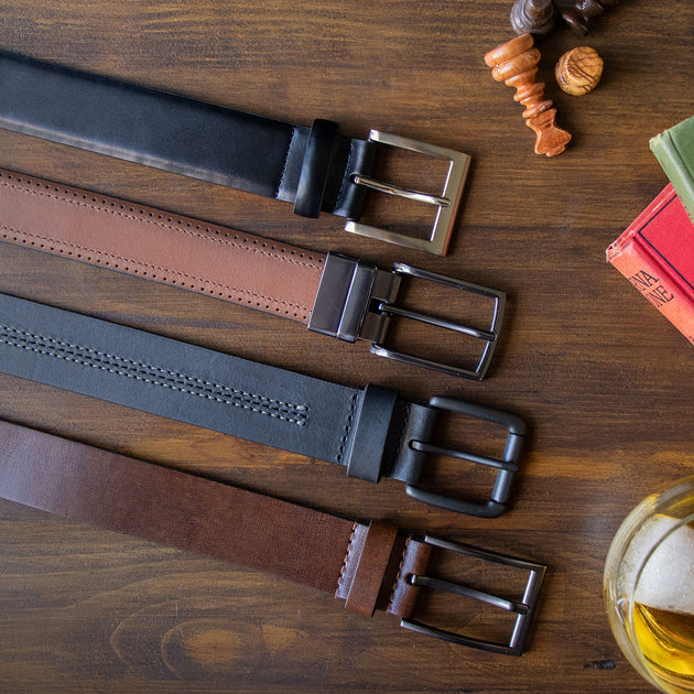 Four leather belts in black and brown tones are arranged diagonally on a dark wooden tabletop, each with a different metal buckle. 